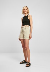Svart avkortet tanktopp, beige høy midje shorts med elastisk liv og knappedetalj, sammen med mørke sandaler, mot en enkel bakgrunn.