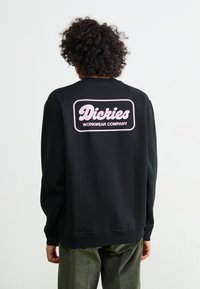 Felpa nera con logo rosa "Dickies Workwear Company" sulla parte posteriore. Realizzata in tessuto morbido con colletto e polsini a costine.