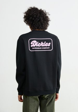 Black sweatshirt featuring pink "Dickies Workwear Company" logo on the back. Made of soft fabric with ribbed collar and cuffs.