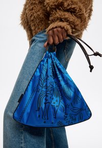 Bolsa de cordón azul con patrones de animales y estrellas en negro, hecha de tela suave, sostenida por una mano con detalles marrones en la correa.