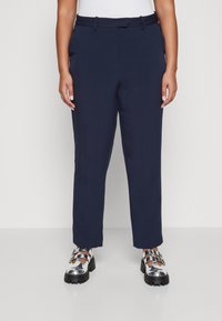 Femme portant un pantalon droit bleu marine et des chaussures à boucle épaisse argentées, se tenant contre un fond blanc.