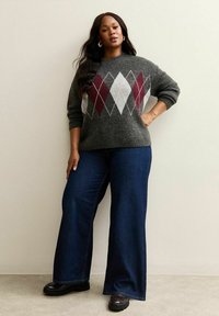 Femme se tenant avec une main sur la hanche, portant un pull en argyle gris, un jean wide-leg bleu foncé et des chaussures noires contre un mur uni.