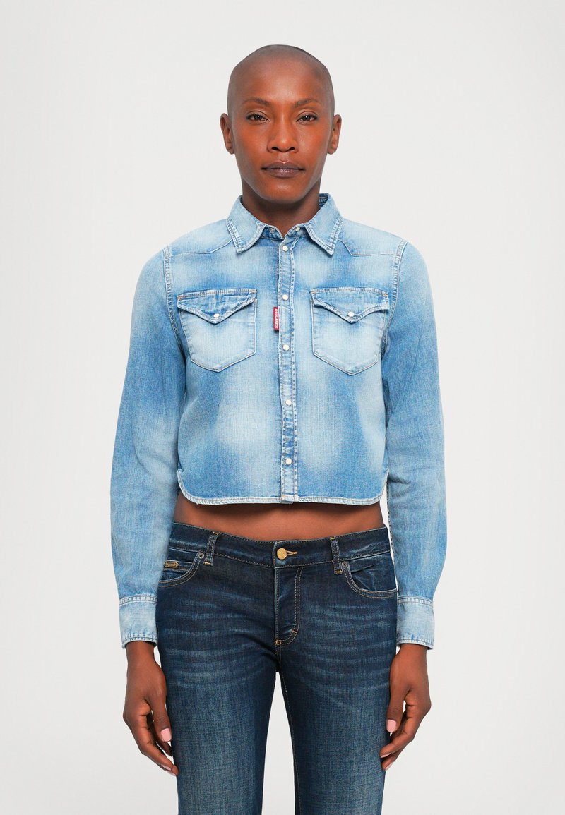 Person wearing a faded light blue cropped denim shirt with two chest pockets and dark blue jeans against a plain white background.