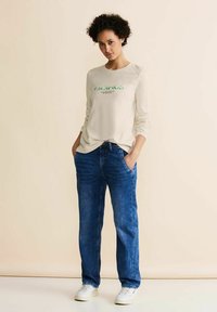 Camicia a maniche lunghe di colore crema con testo verde, abbinata a jeans in denim blu a vestibilità rilassata. Il modello sta in piedi con le mani in tasca, indossando sneakers bianche.