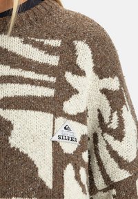 Close-up van een bruin en crèmekleurig gebreid sweater met een bloemenpatroon en een klein driehoekig Quiksilver logo op de borst.