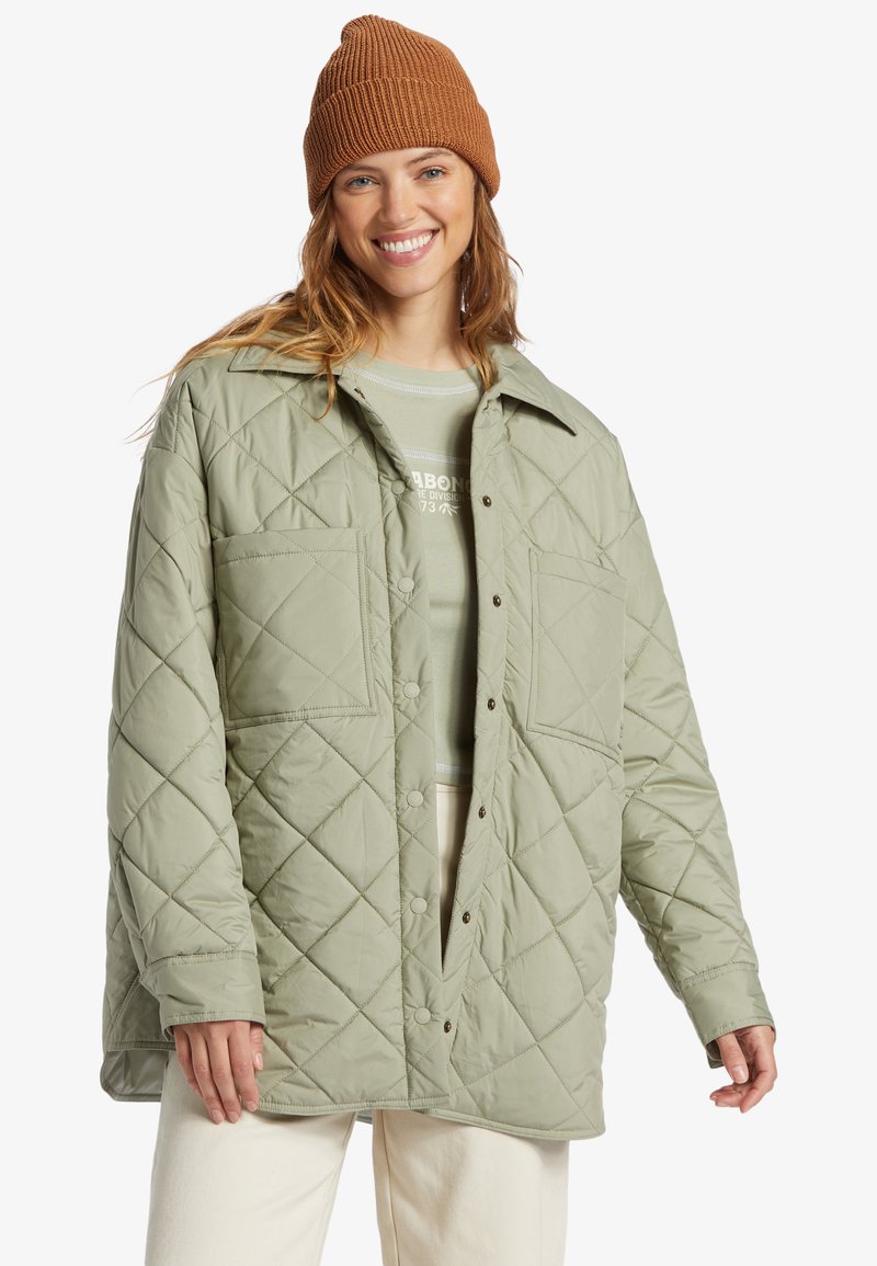 Lys grønn quilted jakke med store lommer, knaplukning og krave. Båret over en grå top. Brun strikket hue på hovedet.