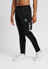 Sorte træningsbukser med en slim fit, elastisk talje og hvidt logo; har hvide chevron-detaljer på siden. Bæret med hvide sneakers.