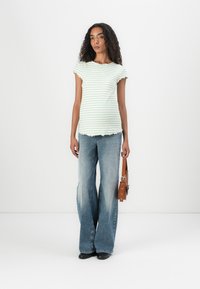 Striped green fitted top with scalloped edges, paired with light blue flared jeans. Model holds a brown shoulder bag, positioned in front of a white background.