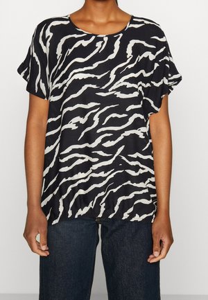 Zwarte loszittende top met korte mouwen en witte onregelmatige zebra-achtige strepen, gedragen met donkerblauwe spijkerbroek.