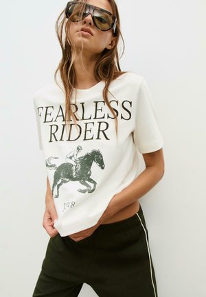 Kvinna som bär överdimensionerade solglasögon och en vit T-shirt med texten "FEARLESS RIDER" samt en grafik av en häst och ryttare, i kombination med svarta byxor.