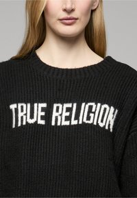 Schwarzer Strickpullover mit den Worten "TRUE RELIGION" in fetten weißen Buchstaben. Die Struktur ist gerippt und hat einen breiten Halsausschnitt.