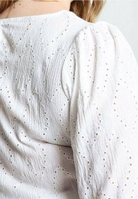 Blouse blanche texturée avec manches bouffantes et petits motifs de découpes en forme de losange portée par une personne vue de dos.