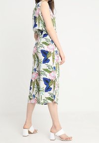 Vestido floral con mariposas azules y flores rosas sobre fondo blanco. Presenta un diseño sin mangas y cintura ceñida. Estilizado con tacones de bloque blancos.