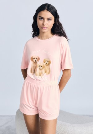 Jeune femme aux longs cheveux foncés portant un ensemble rose à manches courtes et short, orné de trois chiots et des inscriptions "Sweet dream" et "GUESS."