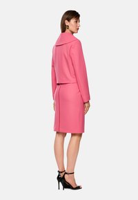 Rosa maßgeschneiderter Anzug mit verkürzter Jacke und knielanger Rock. Glattes Material, großer Kragen, Rückenreißverschluss, kombiniert mit schwarzen, geschnürten High Heels.