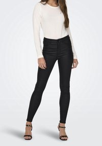 Svarta skinny jeans med hög midja och dragkedjestängning, kombinerat med en långärmad vit topp. Skorna är svarta klackskor med remmar.