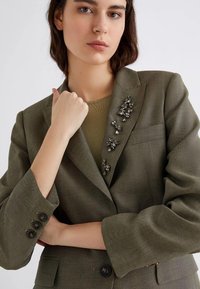 Blazer a spina di pesce verde oliva con spalle strutturate, maniche a due bottoni e un dettaglio a spilla con varie gemme sul risvolto.