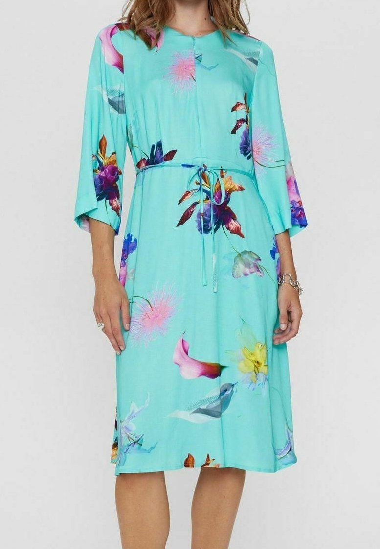 Femme portant une robe turquoise mi-longue aux motifs floraux roses, violets et jaunes, manches trois-quarts, nouée à la taille.