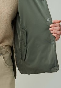 Grüne gepolsterte Weste mit Druckknöpfen und einer Seitentasche. Weiche Innenauskleidung sichtbar, kombiniert mit einem beigen Pullover und hellen Hosen.