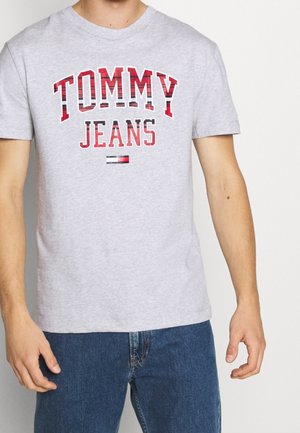 Hombre vistiendo una camiseta ligera de color gris con el texto y logo rojos de "TOMMY JEANS", combinada con unos jeans de mezclilla azules, frente a un fondo neutro.