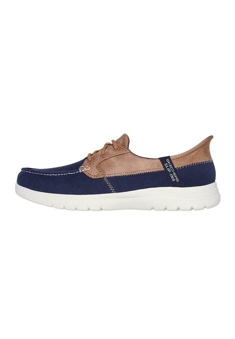 Zapato navy y beige con parte superior de lona y sintético, que presenta una suela blanca, costuras tonales y un diseño sin cordones. Etiqueta de la marca en el lateral.