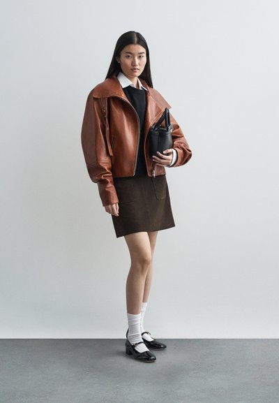 Veste bomber en cuir marron avec un large col, pull noir, jupe marron, chaussettes blanches jusqu'aux genoux et chaussures à talons noires avec des accents.