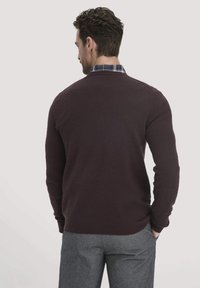 Burgunderfarbenes Strickpullover mit rundem Halsausschnitt, gerippten Bündchen und Saum, über einem karierten Hemd getragen, von hinten betrachtet.