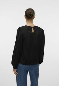 Sort tekstureret bluse med lodrette striber, lange puffede ærmer og en nøglehulldetalje på ryggen, parret med blå lige ben jeans.