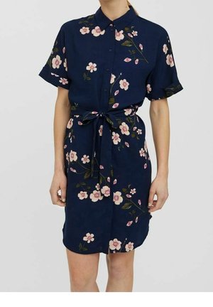 Granatowa sukienka na guziki z krótkimi rękawami, w kwiatowy wzór, z przewiązanym paskiem w talii, noszona przez stojącą osobę z rękami wzdłuż ciała.