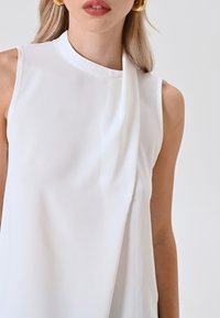 Blusa bianca senza maniche con colletto alto e dettaglio in tessuto drappeggiato su un lato, realizzata in materiale leggero e liscio.