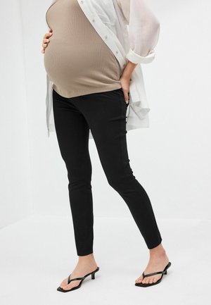 Zwangere persoon draagt een beige top, zwarte strakke broek, doorschijnend wit overhemd met knopen en zwarte sandalen met hak, staat met één hand op de buik.