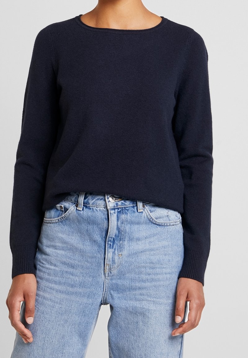 Pull en maille bleu marine à col rond et manches longues, associé à un jeans en denim bleu clair. Le pull présente des poignets et un ourlet côtelés.