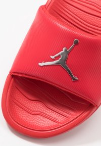 Punaiset urheilusandaalit, joissa on teksturoitu hihna ja kohotettu pohja, jossa on hopea Jumpman-logo. Kevyet ja muotoiltu sisäpohja.