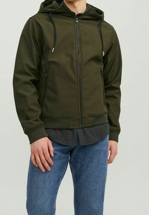 Mężczyzna noszący oliwkowo zieloną kurtkę z kapturem na zamek błyskawiczny ponad ciemną, kraciastą koszulą i niebieskie dżinsy, stojący na jednolitym jasnym tle.