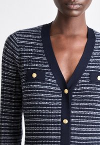 Námořnický svetr s bílými pruhy, výstřihem do V, zlatými knoflíky a dvěma předními kapsami. Texturovaná tkanina s kontrastními vroubkovanými okraji.