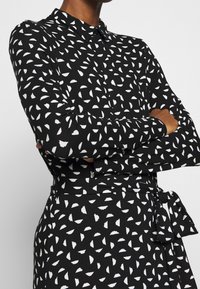 Svart långärmad skjortklänning med ett vitt geometriskt mönster, krage och ett band i midjan för justering.