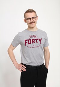 Camiseta de algodón gris con "Cumplir CUARENTA es ejercicio suficiente" en letras color marrón, combinada con pantalones negros y un cinturón negro, de corte ajustado.