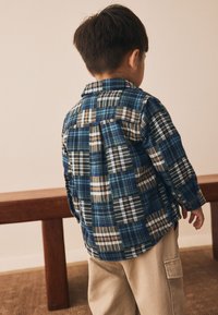 Blå og brunternet flannel-skjorte med lange ærmer, der har forskellige firkantede mønstre; kombineret med beige cargobukser. Set bagfra.