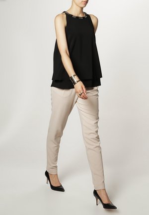 Top nero senza maniche con un design a strati e un collo decorato, abbinato a pantaloni beige chiaro slim e scarpe nere col tacco alto.