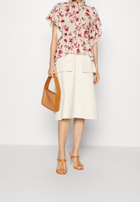Donna che indossa una blusa floreale con collo alto, una gonna beige fino al ginocchio con tasche, tacchi arancioni a strappo e tiene in mano una borsa intrecciata marrone.