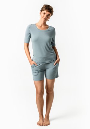 Woman stands barefoot with hands in pockets wearing light blue short-sleeve top and matching shorts against plain white background.