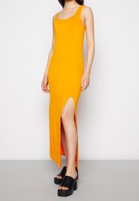 Długa, dopasowana sukienka w jaskrawym pomarańczowym materiale z dekoltem w kształcie łódki i rozcięciem do uda. Połączona z czarnymi sandałami na platformie.