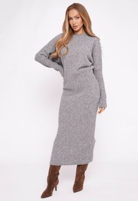 Kobieta w długiej szarej prążkowanej dzianinowej sukience i brązowych zamszowych botkach na wysokim obcasie, pozująca z jedną ręką na biodrze na białym tle.