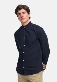 Chemise bleu marine à boutons, en coton, avec un col classique, des manches longues et un petit logo sur le côté gauche de la poitrine. Tissu texturé.