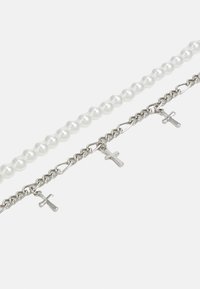 Tres pulseras: una de perlas blancas, otra de cadena de plata con colgantes de cruz, y una tercera de eslabones de plata entrelazados.
