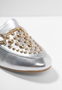 Chaussure en cuir argenté à enfiler, ornée de clous et accents métalliques dorés, avec un bout arrondi, une texture lisse et un profil élégant.