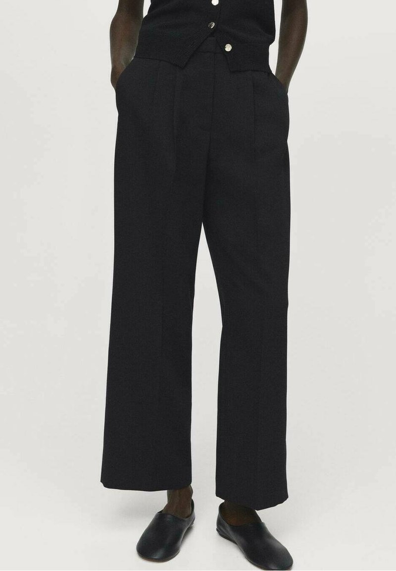 Personne portant un pantalon noir taille haute à jambes larges avec des poches latérales, associé à des chaussures noires à enfiler, sur un fond uni.