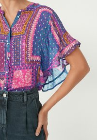 Blusa colorata a fantasia con maniche corte a sbuffo, caratterizzata da design geometrici nelle tonalità di rosa, viola e blu; tessuto leggero.