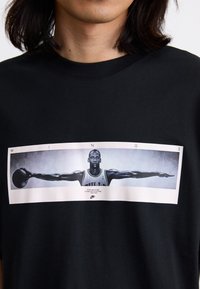 Sort bomulds T-shirt med et stort hvidt kantet grafisk billede af en basketballspiller med udstrakte arme, tekst under i en lys skrifttype.