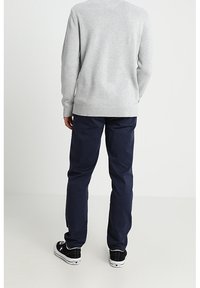 Uomo che indossa un maglione grigio chiaro, pantaloni blu navy e sneaker nere, in piedi rivolto di spalle su uno sfondo bianco semplice.